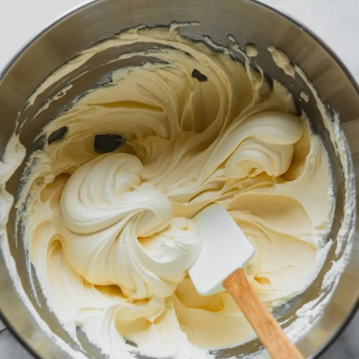 Fluffy cream cheese mousse whipped in a stainless steel mixing bowl with a spatula, showcasing smooth, swirled peaks of the pale yellow mixture.