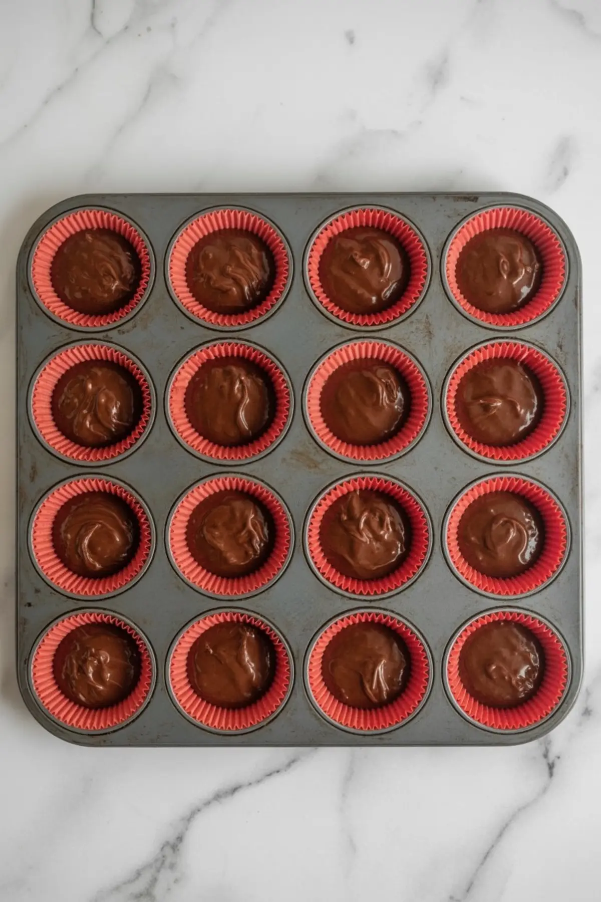 Muffin tin filled with red cupcake liners, each holding a scoop of chocolate cupcake batter, placed on a white marble countertop, prepped for baking.