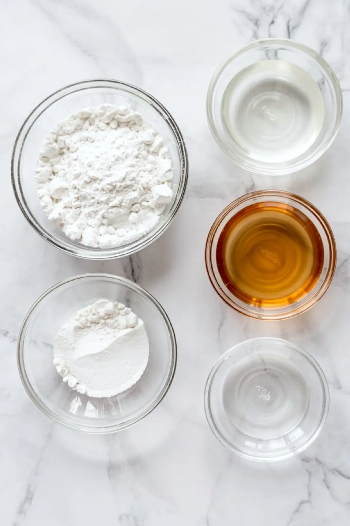 Glass bowls on a marble countertop showing measured ingredients for royal icing, including powdered sugar, cornstarch, meringue powder, vanilla extract, and water.