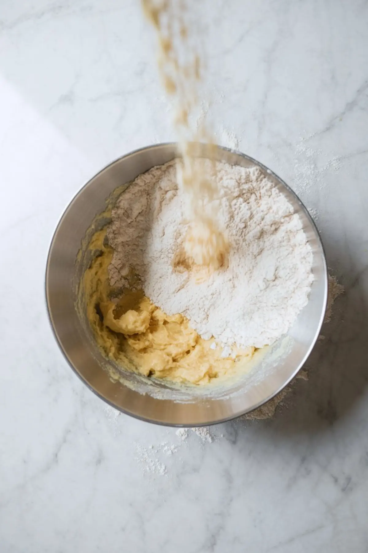 Flour being added to a bowl of creamed butter and sugar on a marble countertop during cookie dough mixing.