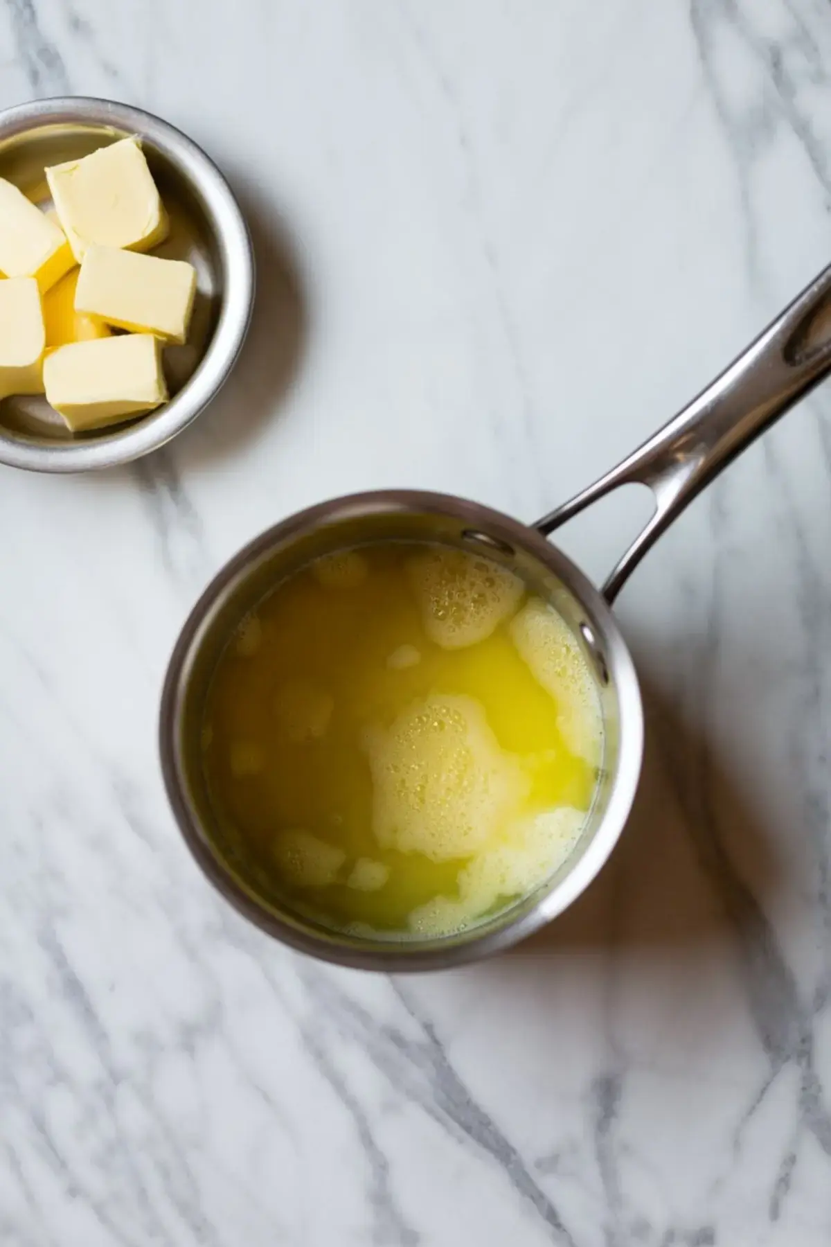 A metal saucepan with melted butter and a bowl of cubed butter set on a marble countertop, ready for baking preparation.