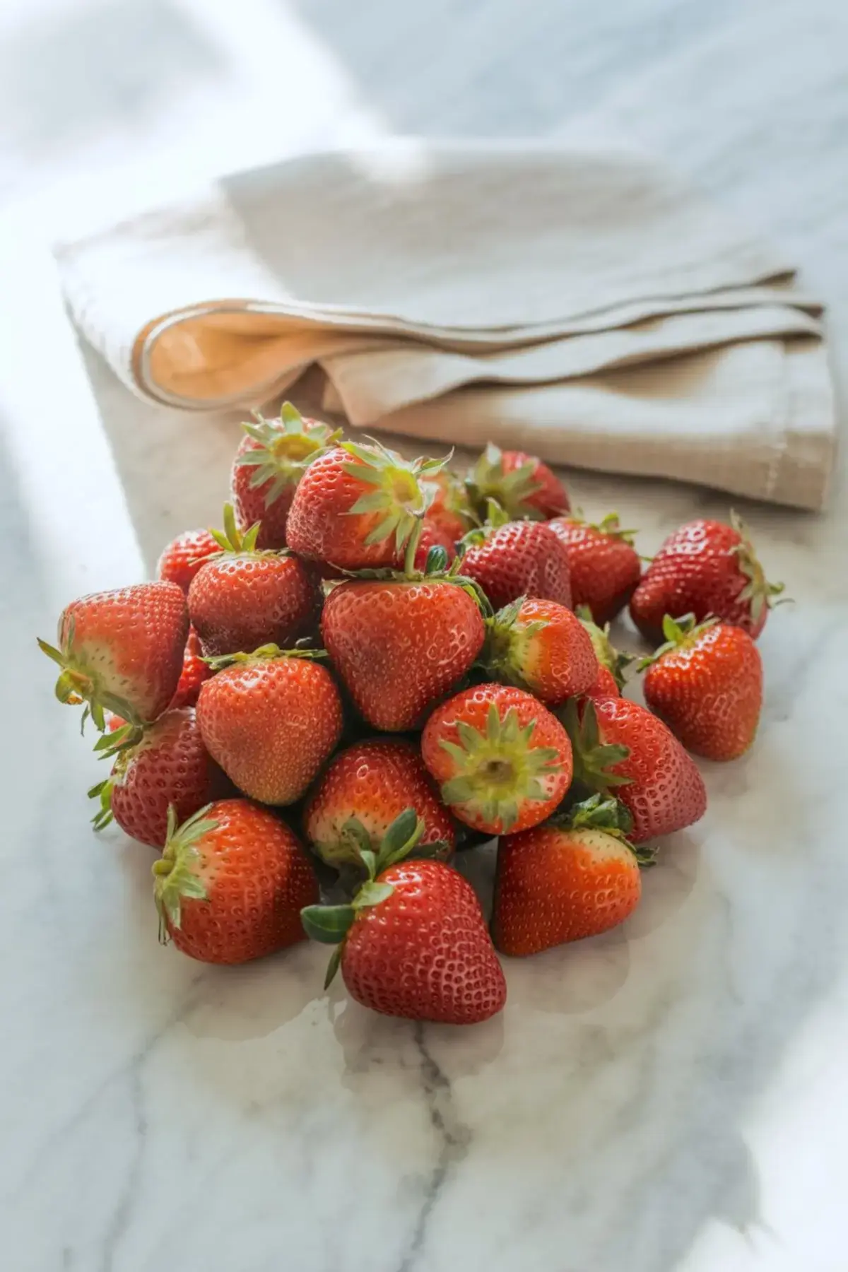 A fresh pile of red strawberries with green tops arranged casually on a marble counter next to a folded beige kitchen towel in natural light.