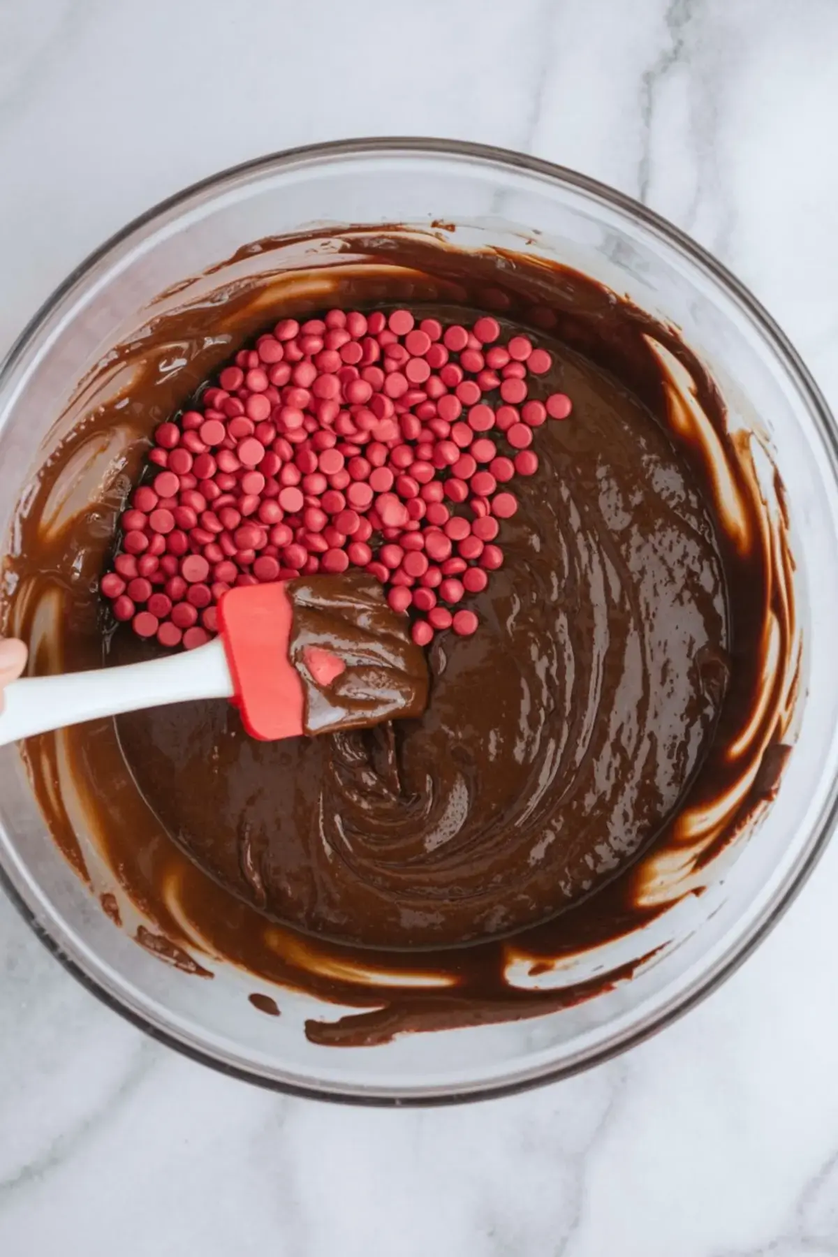 A glass bowl filled with thick chocolate brownie batter being mixed with red candy-coated chocolate chips using a red and white spatula on a marble surface.