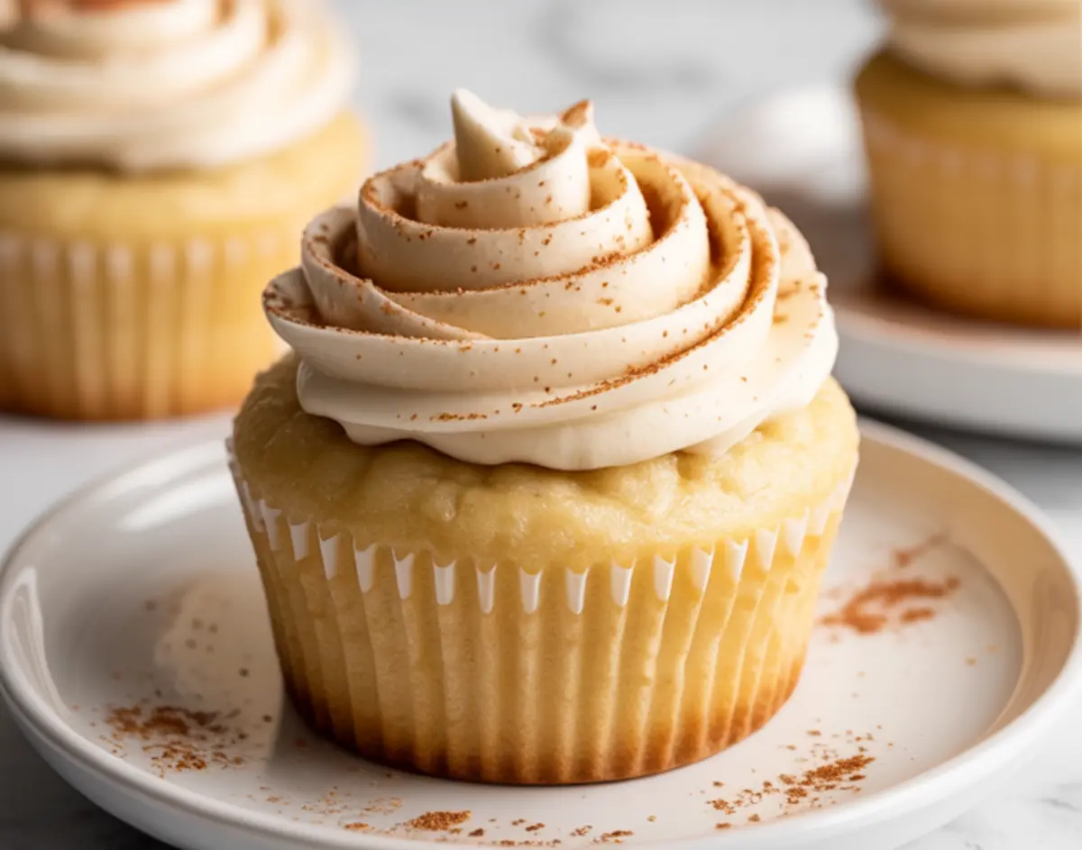 Side view of a single snickerdoodle cupcake with cinnamon buttercream frosting piped in tall swirls, placed on a white dessert plate.