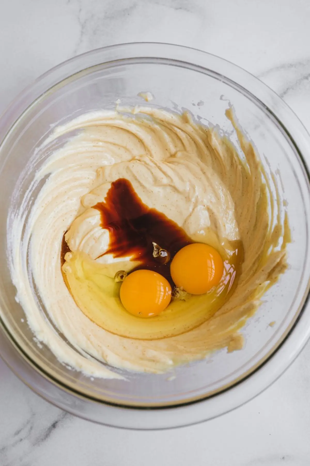 Glass mixing bowl filled with vanilla cake batter, two raw eggs, and a splash of vanilla extract on top, placed on a marble surface.