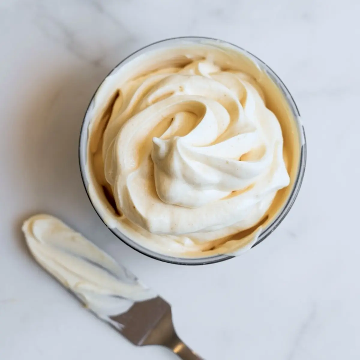 A bowl of fluffy whipped frosting with a smooth swirl on top, placed on a marble surface beside an offset spatula.
