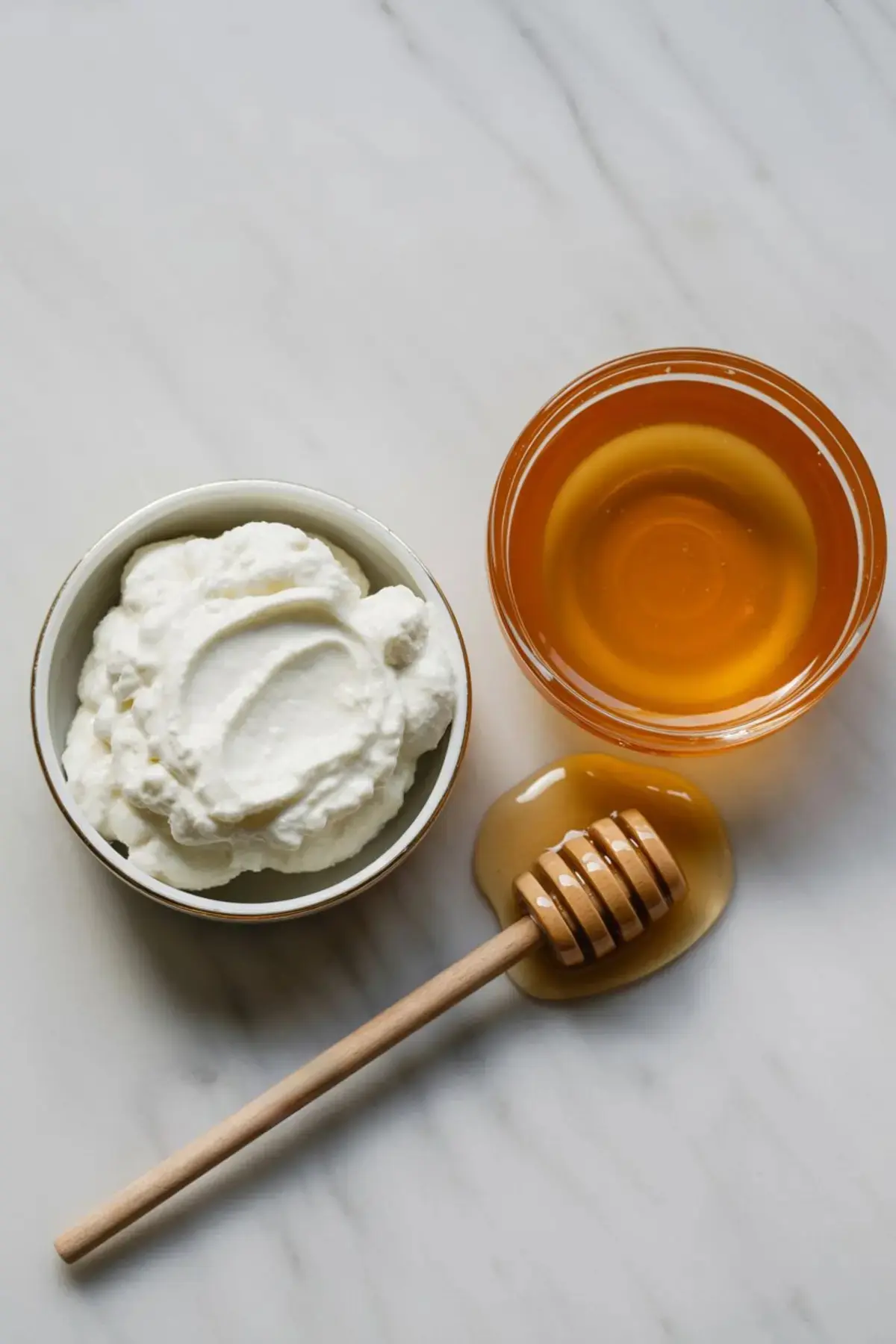 Bowl of ricotta cheese and a glass of honey with a wooden honey dipper resting on a white marble surface.