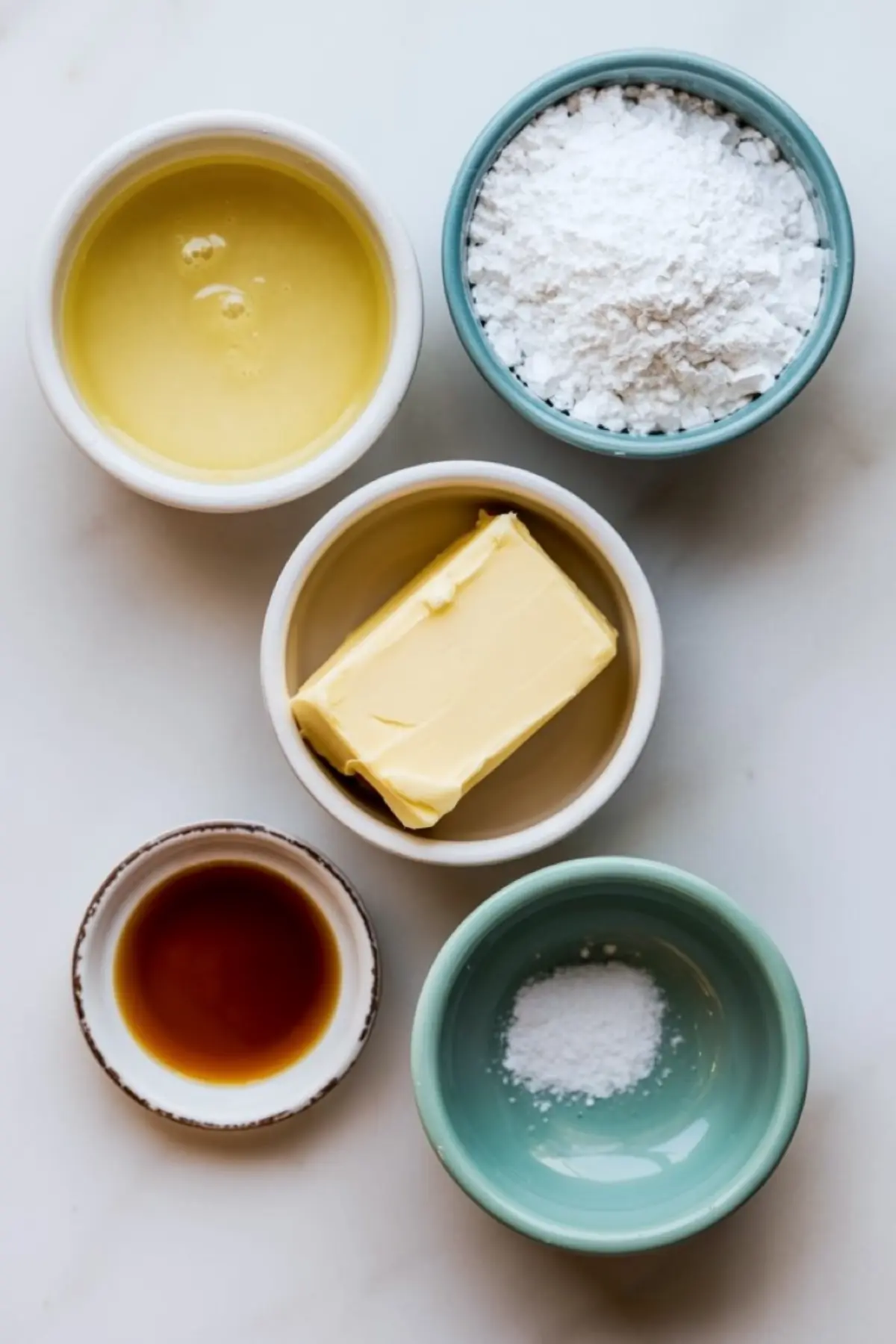 Flat lay of vegan frosting ingredients in small bowls, including powdered sugar, dairy-free butter, vanilla extract, plant milk, and salt, arranged on a white marble background.
