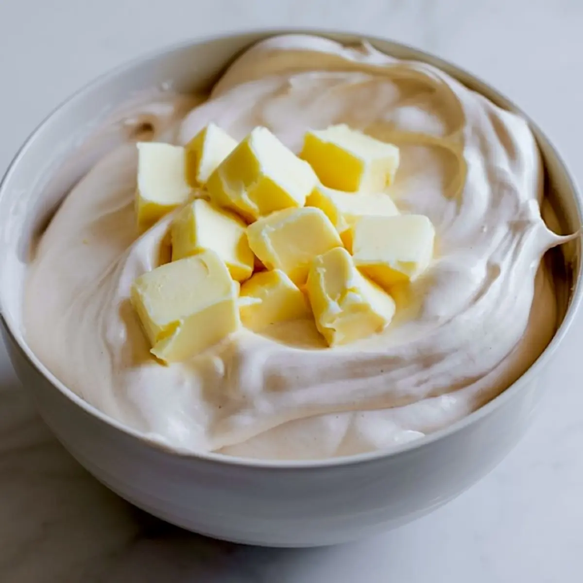 Smooth vegan frosting with added cubes of dairy-free butter in a white bowl on a marble surface, ready for mixing.
