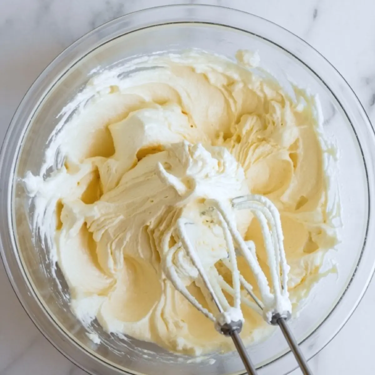 Glass bowl filled with creamy whipped mascarpone mixture, freshly blended with electric beaters on a marble surface.