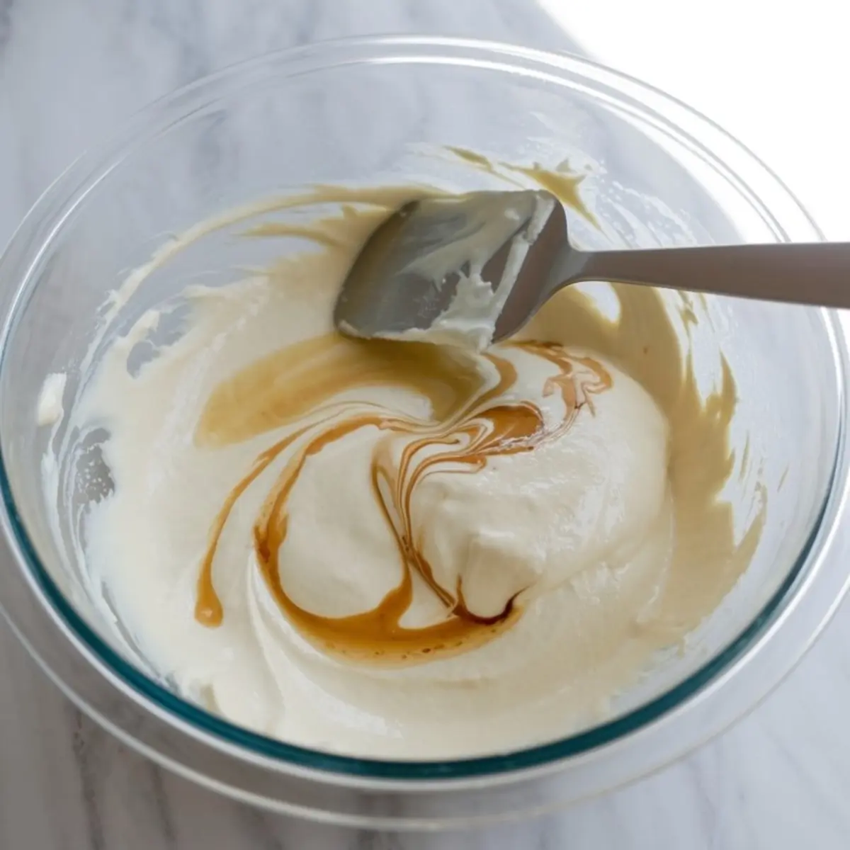 Creamy mascarpone mixture in a glass bowl being swirled with golden vanilla extract using a spatula on a light marble surface.