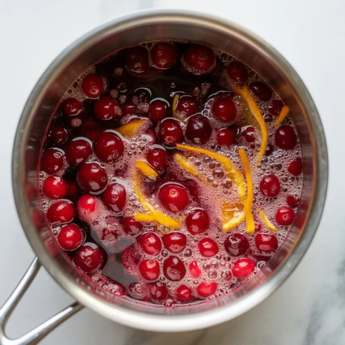 Pot of bubbling cranberry sauce with whole cranberries and strips of orange zest simmering in liquid on a marble background.