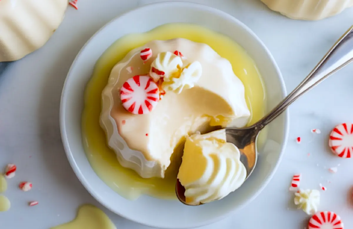 Close-up of creamy white chocolate peppermint panna cotta on a white plate, garnished with crushed peppermint candy and whipped cream, with a spoonful removed to reveal smooth texture.
