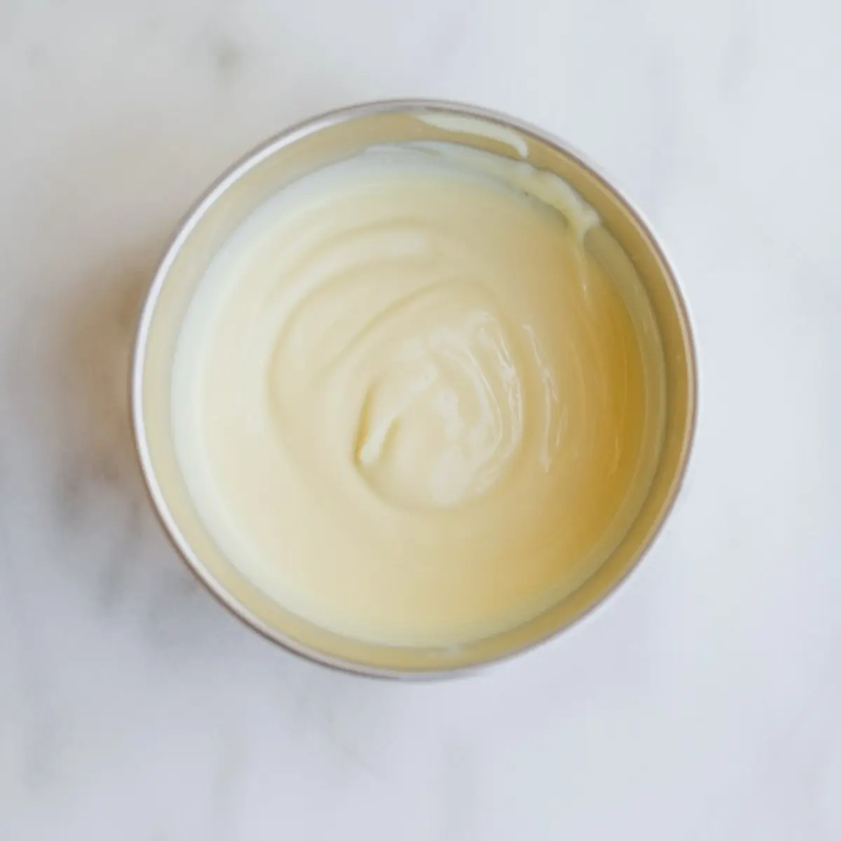 Smooth white chocolate and marshmallow mixture swirled in a metal bowl on a white marble surface, showing a creamy base for rice krispie treats.
