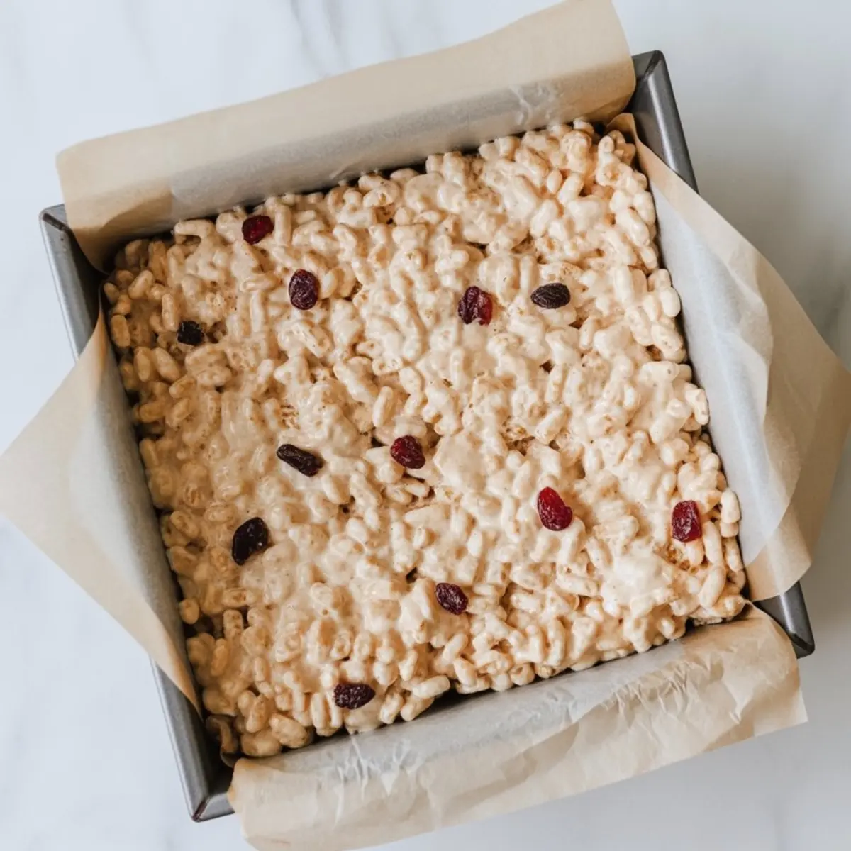 Pressed rice krispie treat mixture with dried cranberries in a parchment-lined square baking pan, ready to be sliced into bars.
