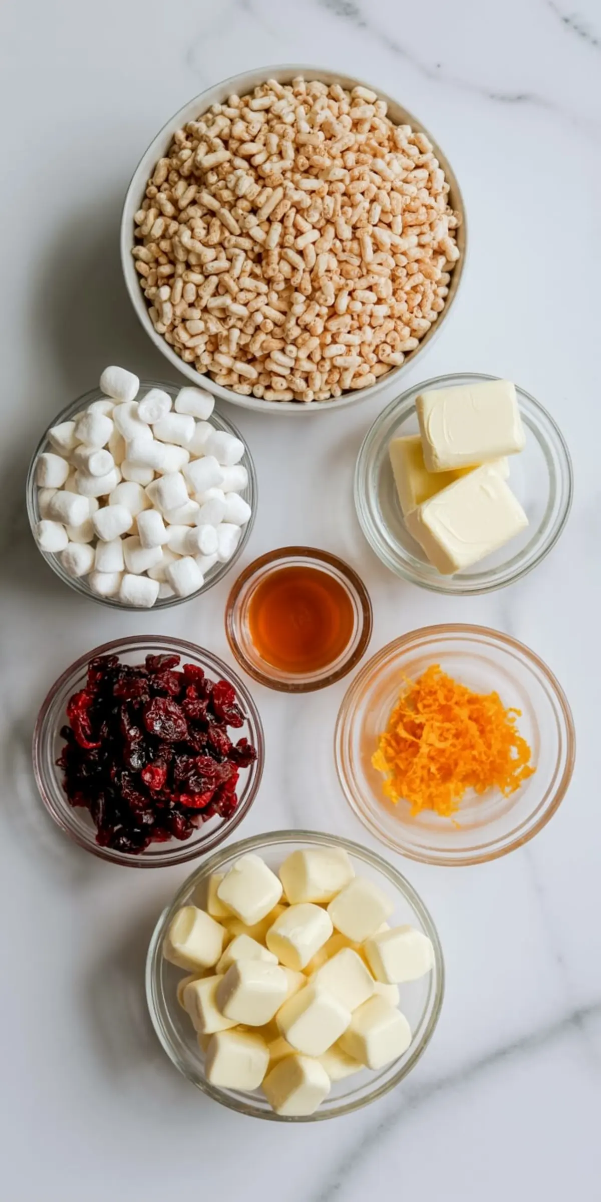 Overhead view of ingredients for white chocolate cranberry rice krispie treats, including puffed rice cereal, mini marshmallows, butter, dried cranberries, vanilla extract, and orange zest in glass bowls on a white marble background.

