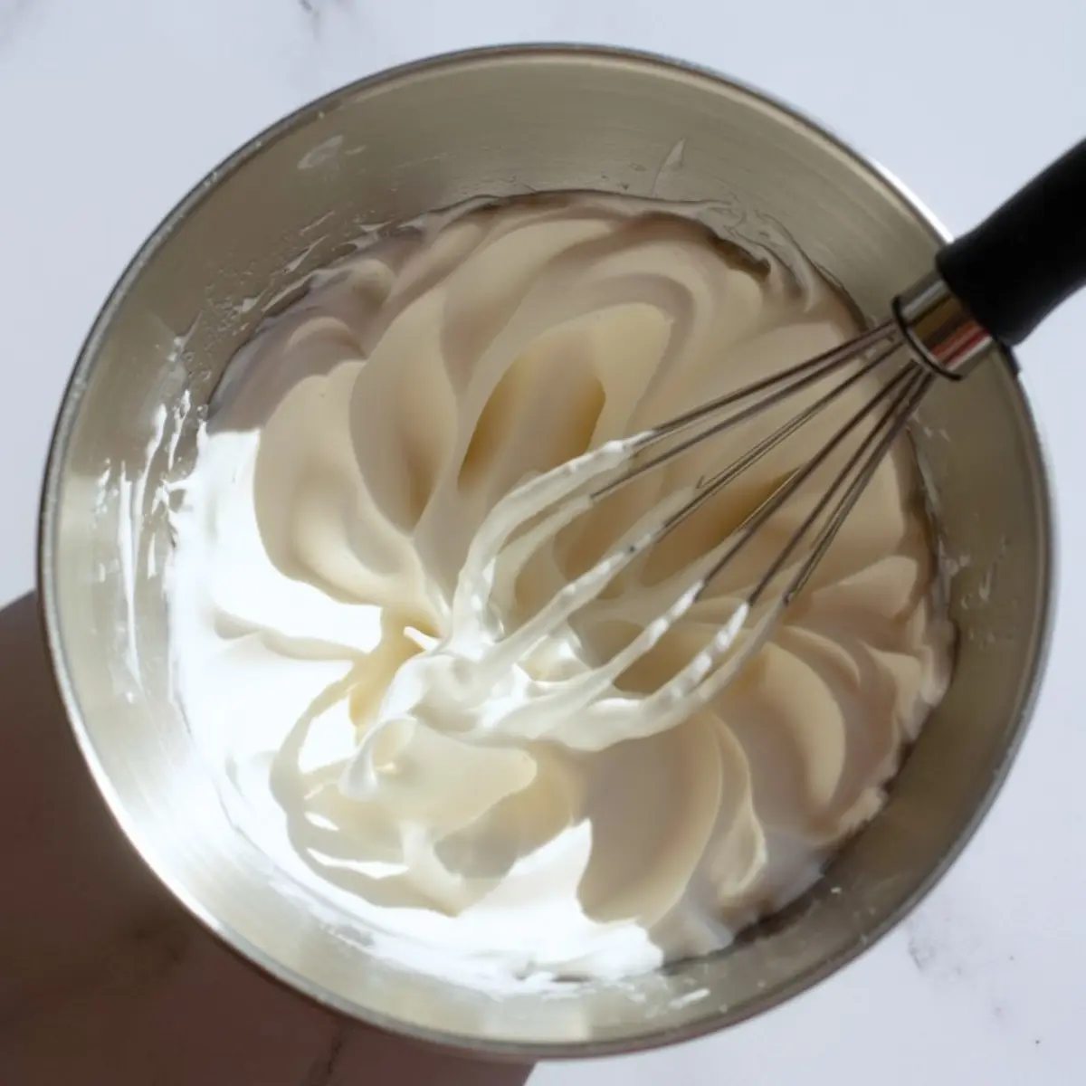 A metal mixing bowl filled with freshly whipped cream, forming soft peaks, with a whisk resting inside.

