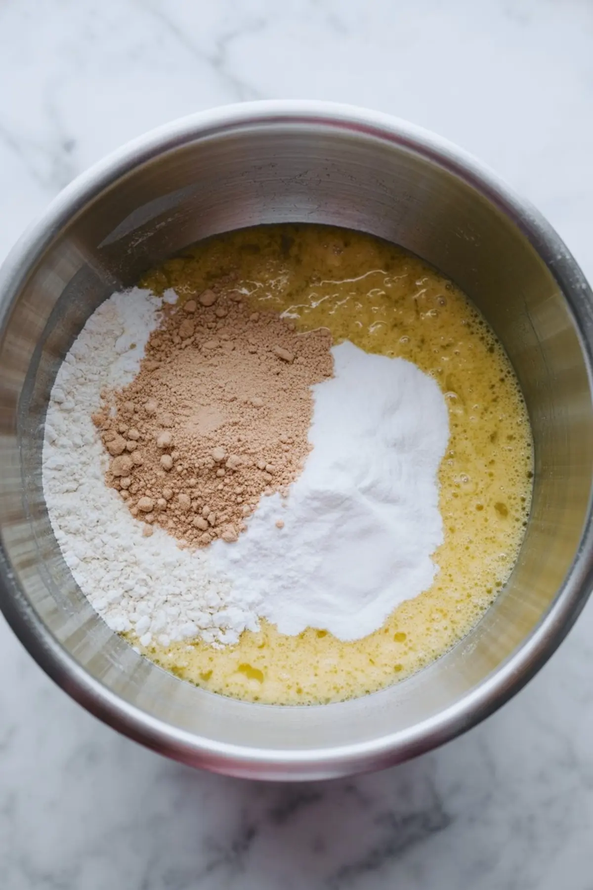 A mixing bowl filled with wet and dry baking ingredients including flour, cocoa powder, and baking soda poured over a whisked yellow batter on a marble countertop.
