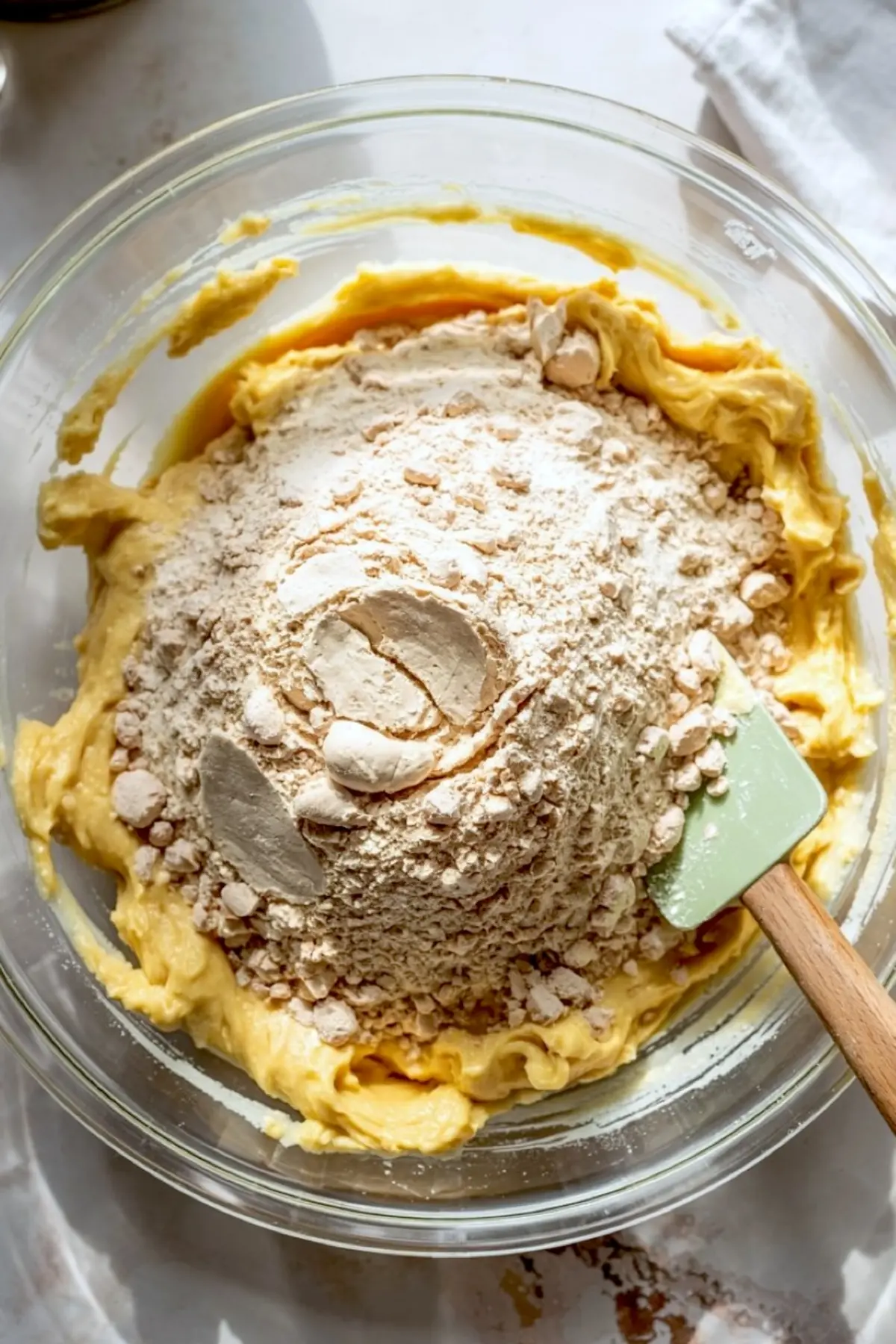 Flour added to a smooth cookie dough base in a glass bowl with a green spatula, showing the early stage of mixing dry and wet ingredients.
