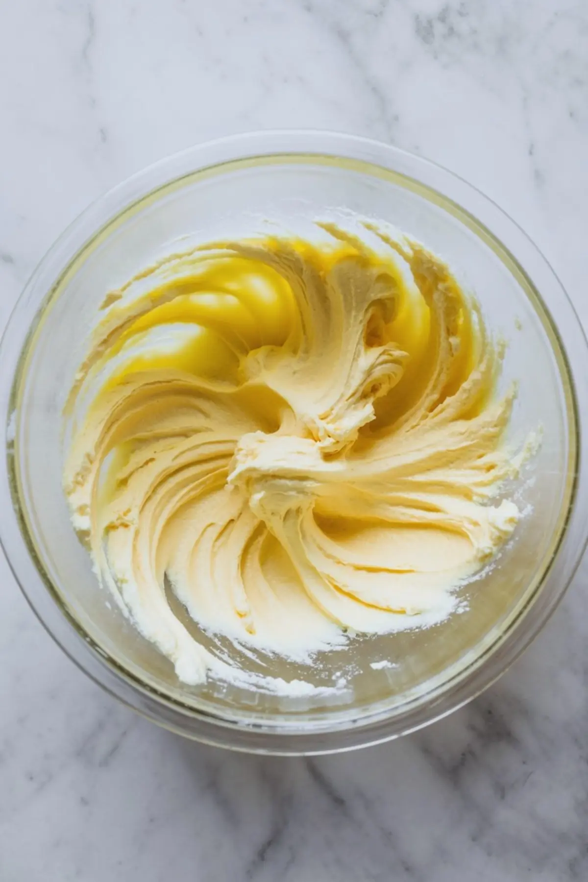 Smooth butter and sugar mixture in a clear glass bowl on a marble surface, showing creamy swirls created during mixing.

