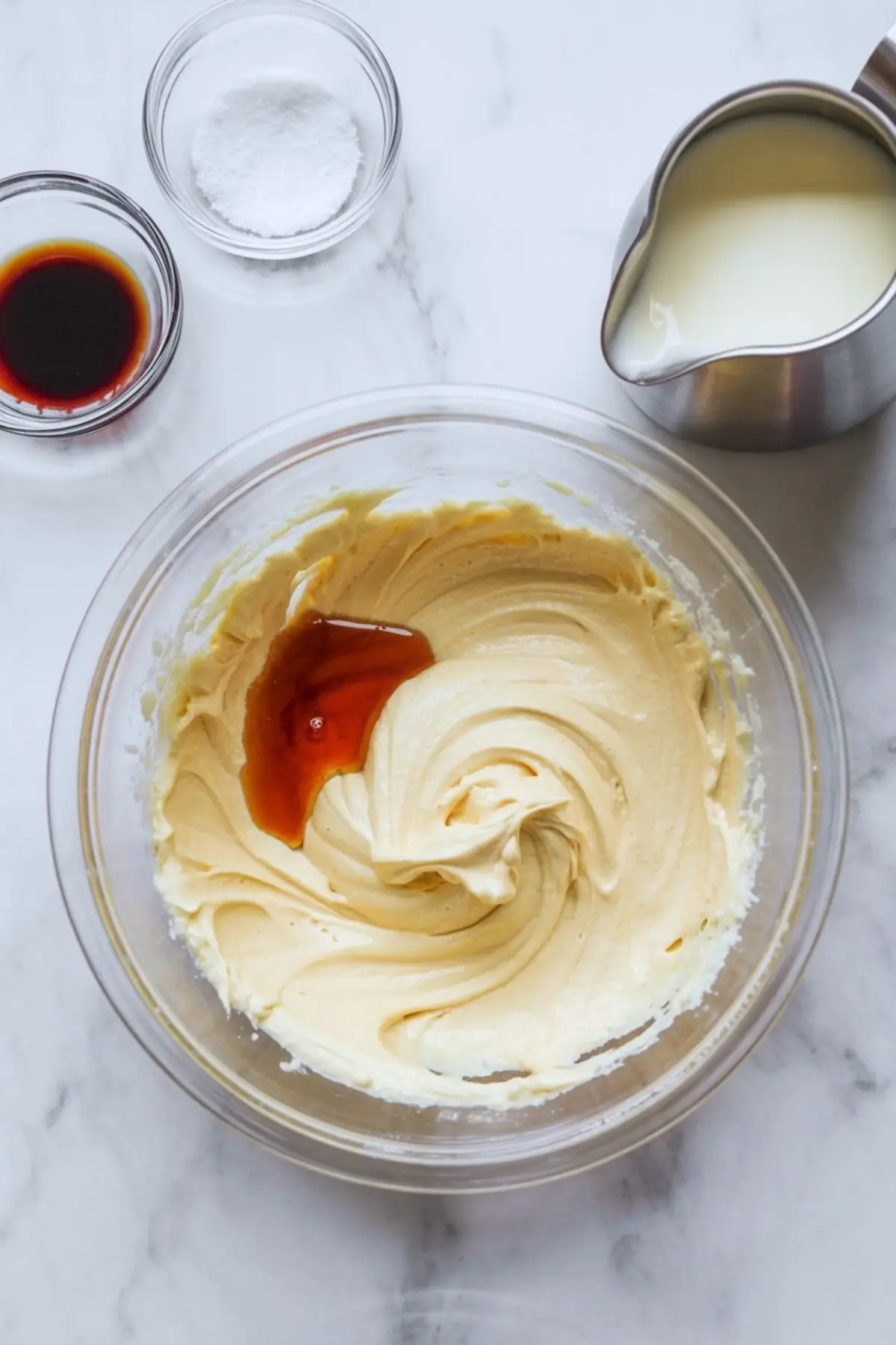 Creamy butter mixture in a glass bowl with vanilla extract poured on top, surrounded by small bowls of sugar, salt, and milk on a marble counter.
