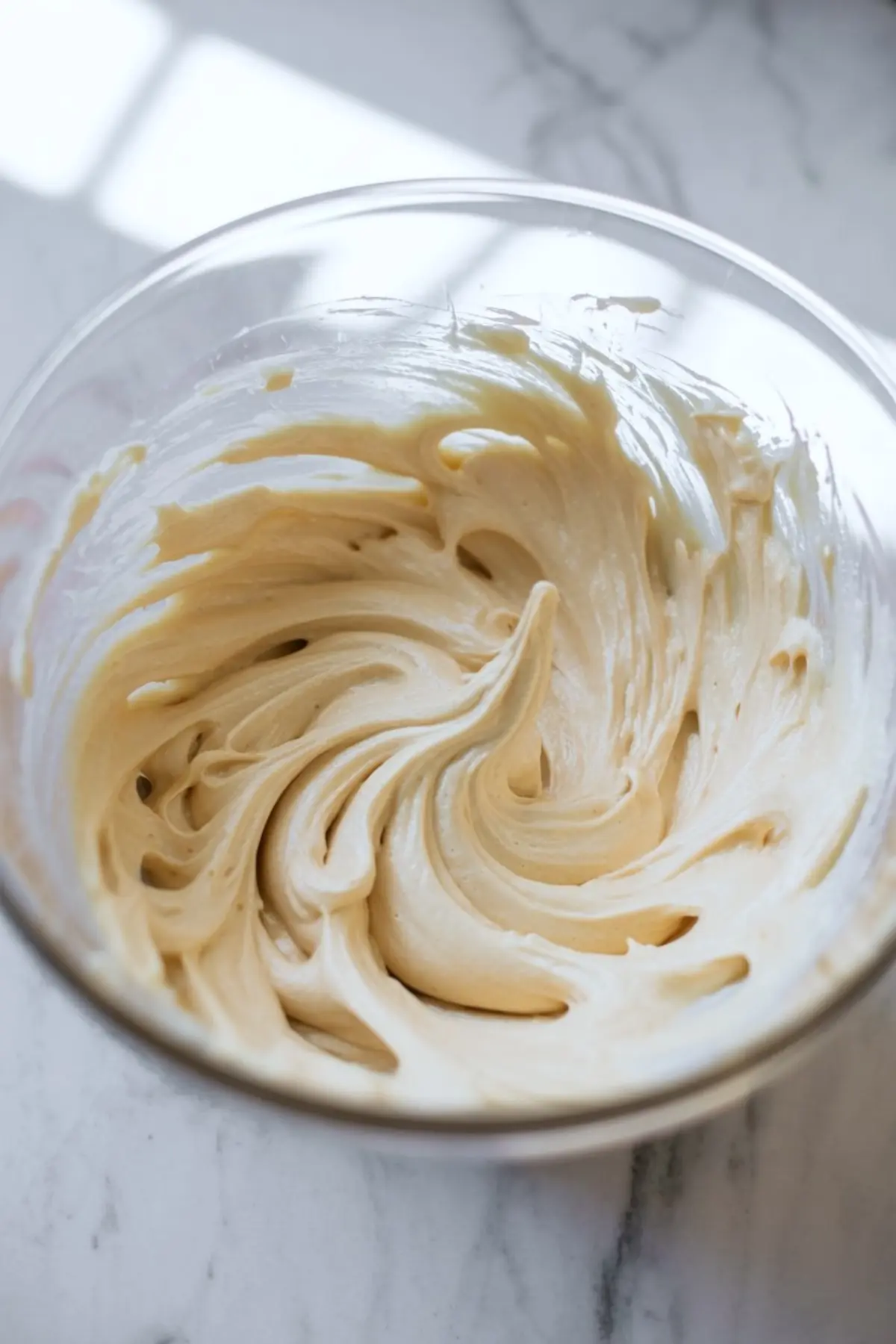 Light and fluffy buttercream mixture in a glass bowl with soft peaks and smooth swirls created by thorough mixing.
