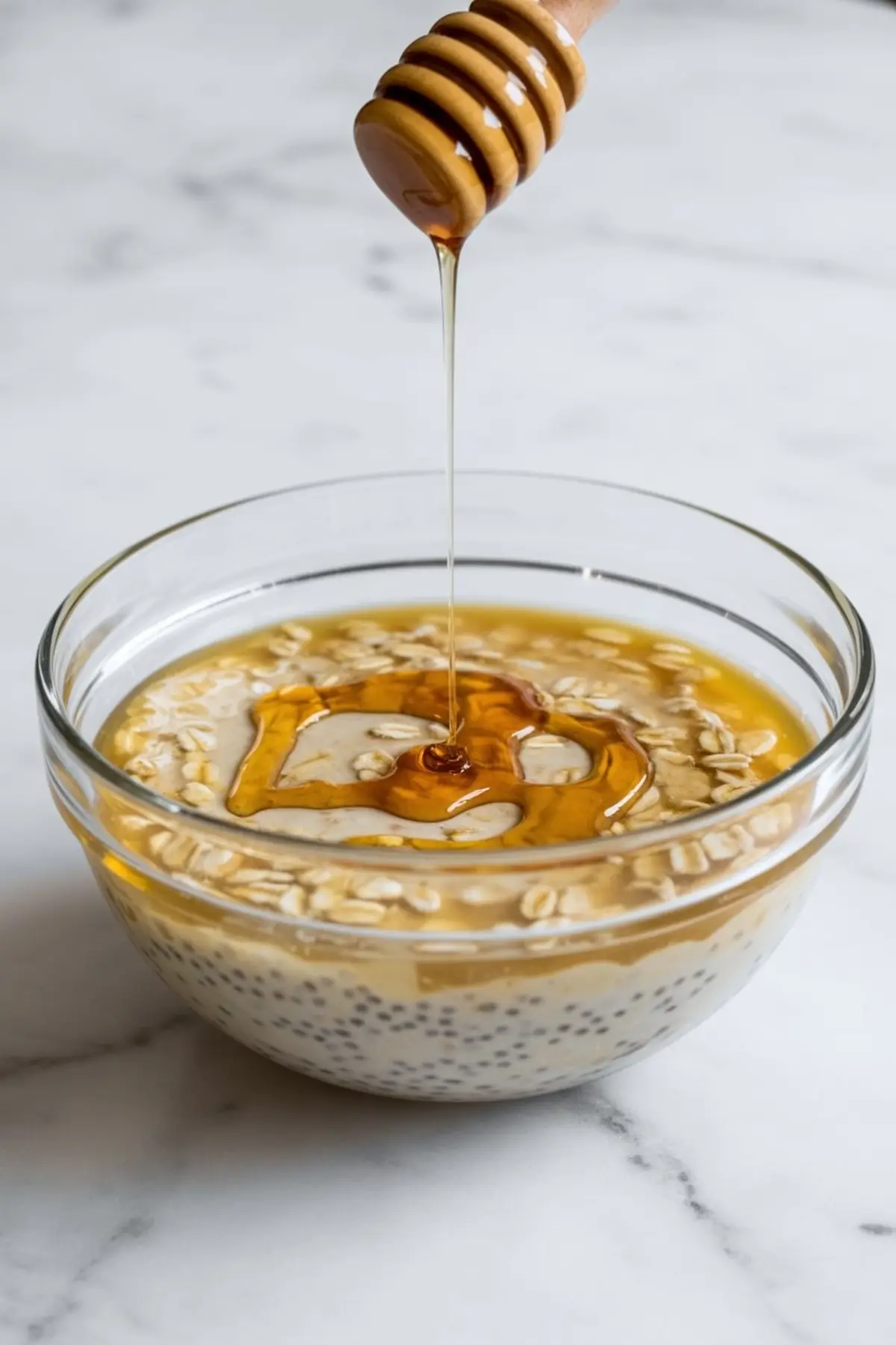 Honey being drizzled from a wooden dipper into a glass bowl of overnight oats made with chia seeds and rolled oats on a marble background.