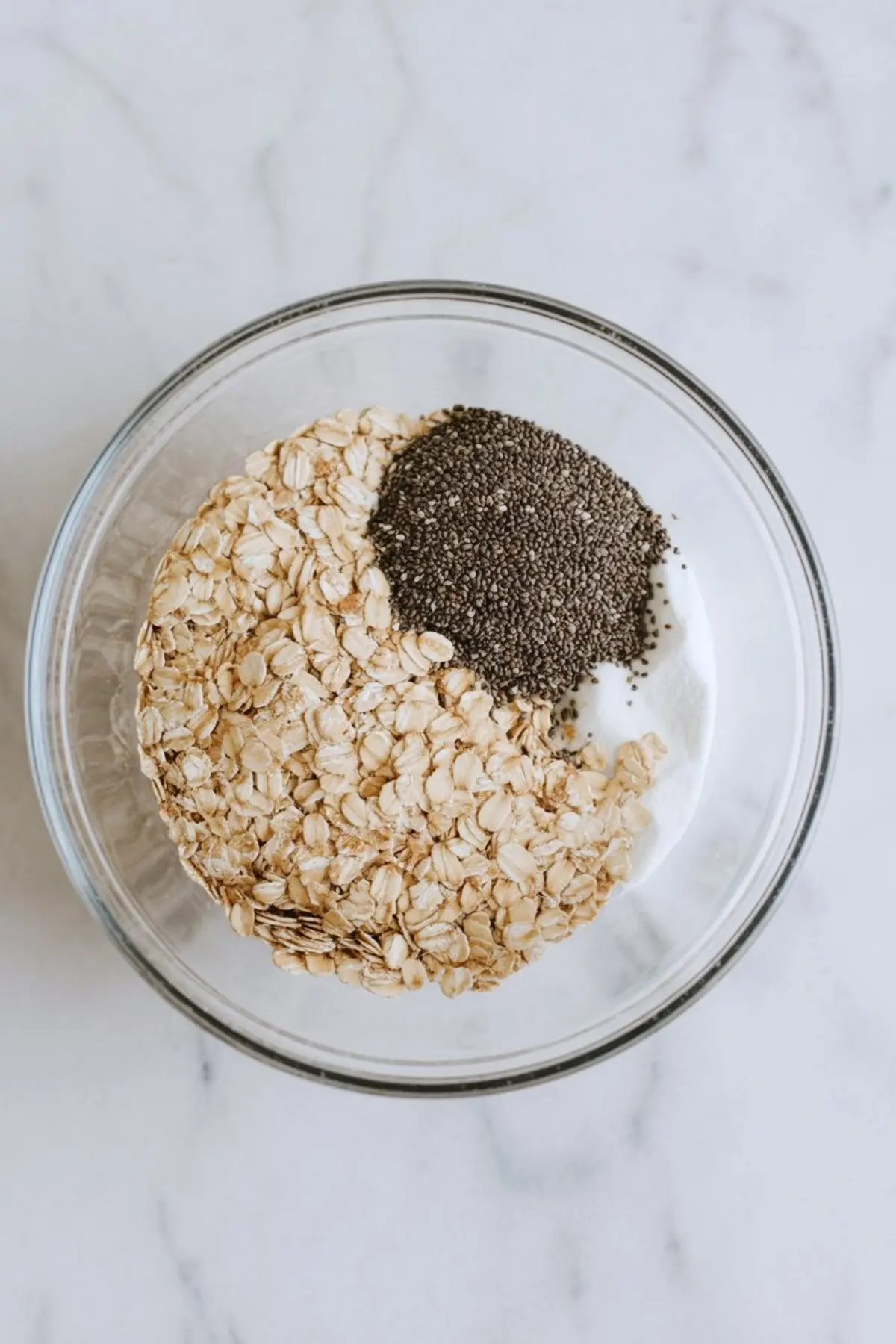 Dry rolled oats and chia seeds layered over yogurt in a glass mixing bowl on a light marble surface, ready to be stirred for overnight oats.