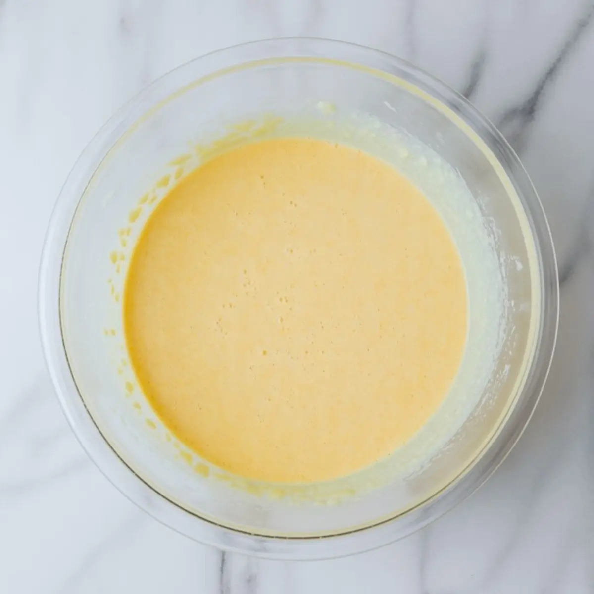 Close-up of a glass mixing bowl filled with smooth, golden yellow French toast custard made from eggs, milk, and sugar on a marble surface.