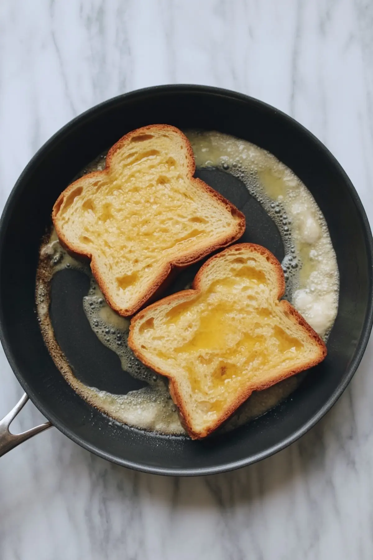 Two slices of brioche bread frying in bubbling butter in a nonstick skillet, golden and soaking up the custard mixture.
