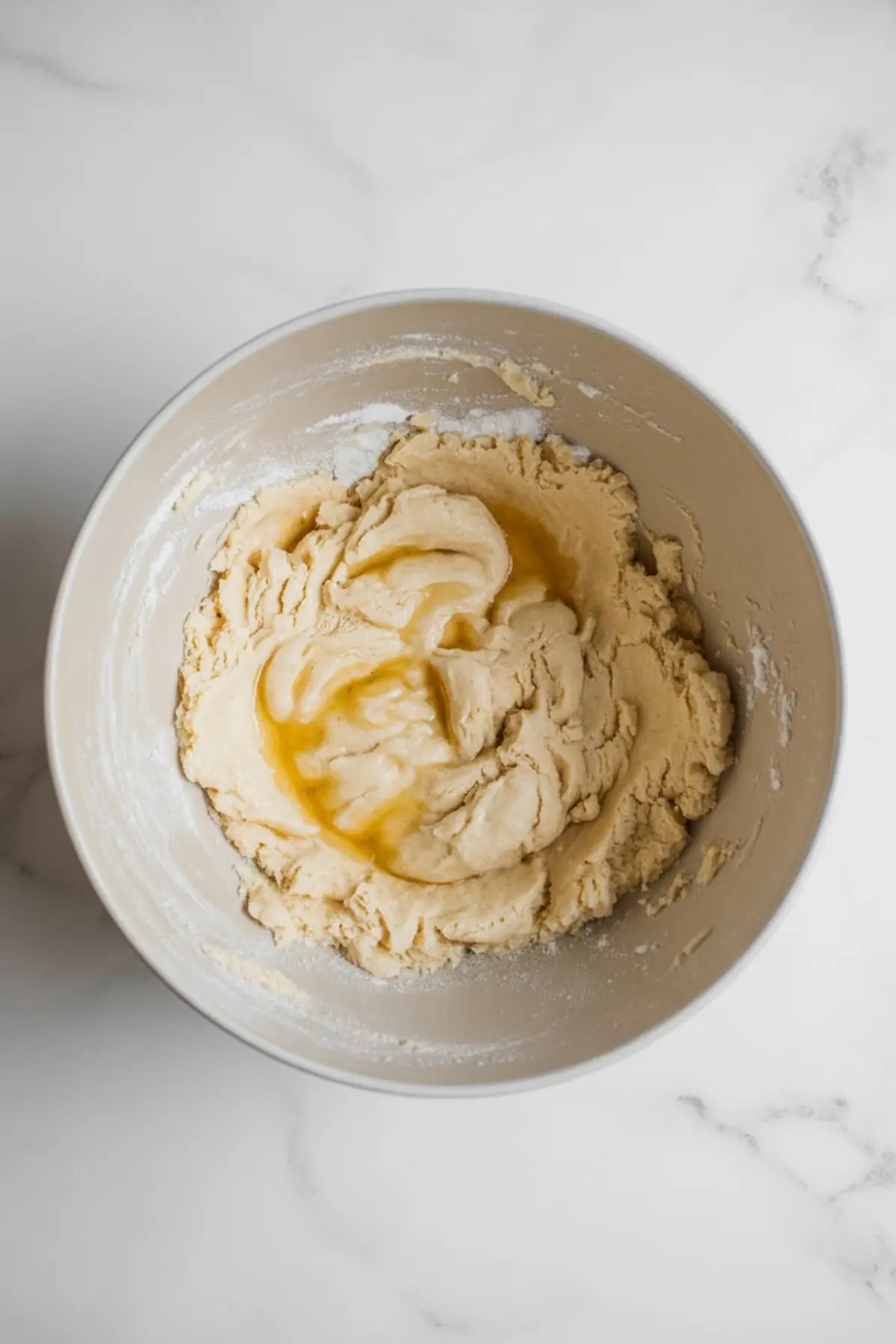 Thick cookie dough mixed with vanilla extract in a ceramic mixing bowl, sitting on a white marble countertop with flour residue around the edge.
