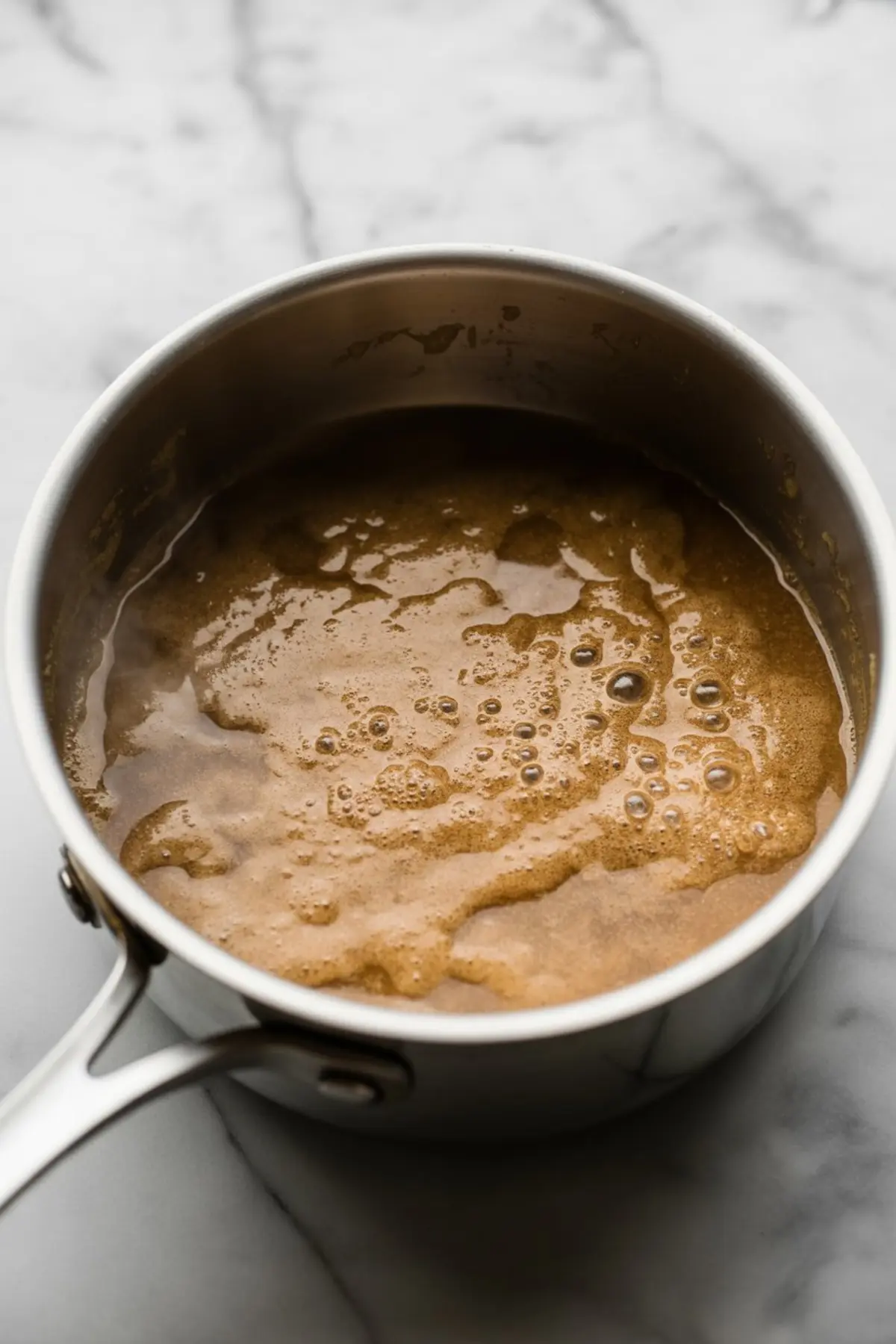 Simmering brown butter mixture with bubbling surface in a stainless steel saucepan placed on a marble counter, showing caramelization in progress.
