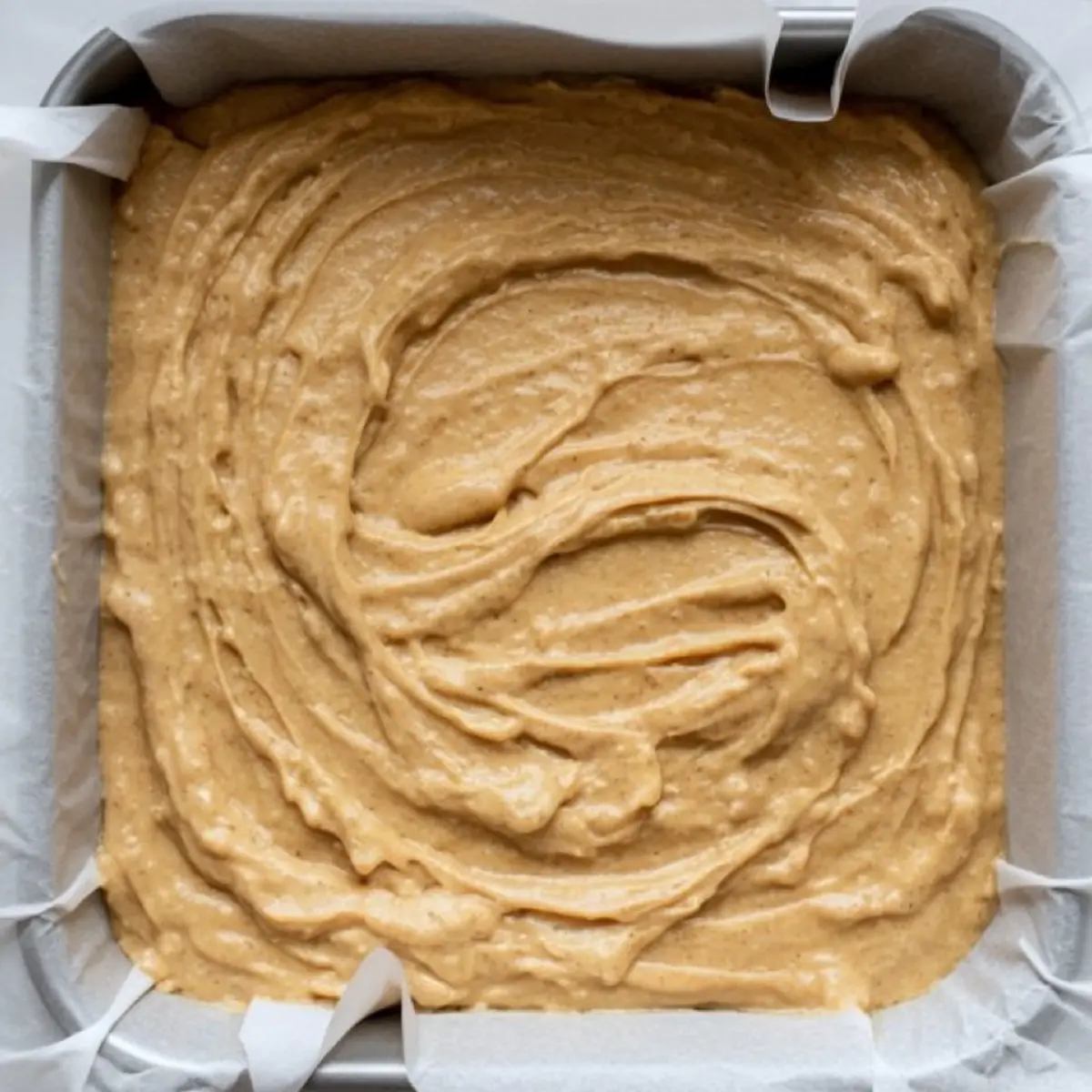 Smooth blondie batter spreads evenly in a parchment-lined square baking pan, ready to be baked for a dense, chewy texture.
