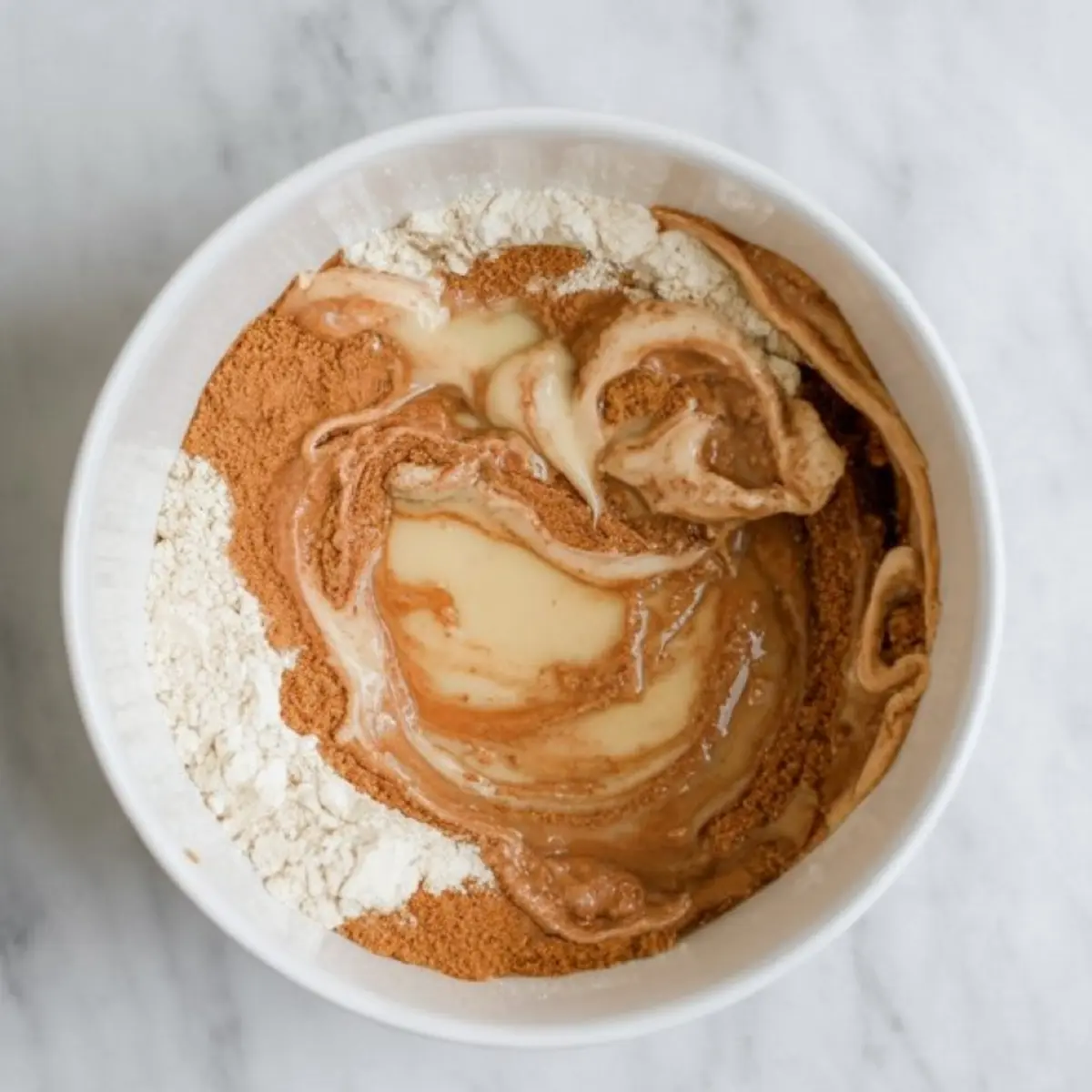 White bowl containing a mixture of flour, cinnamon, and sweetened condensed milk starting to blend into a thick batter.