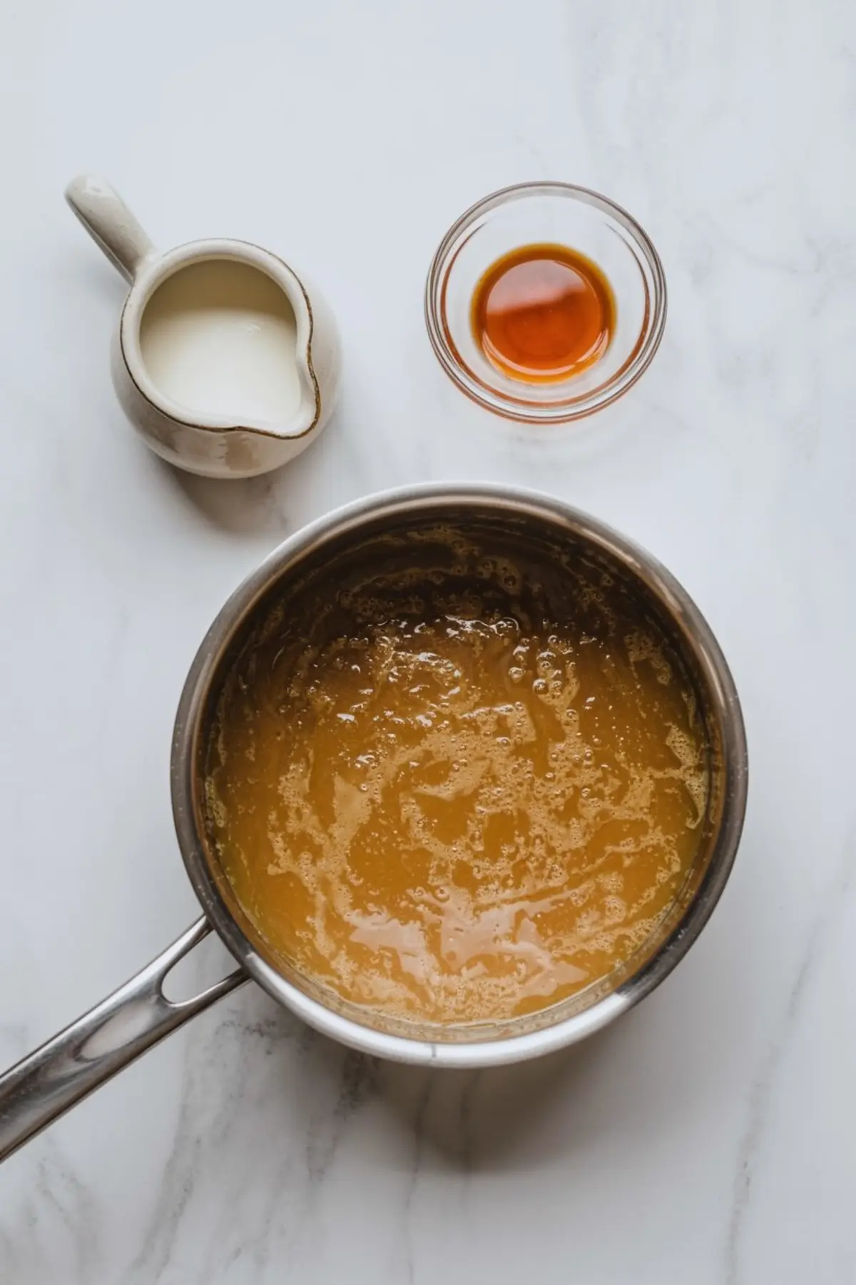 Caramel mixture bubbling in a saucepan with a pitcher of cream and a bowl of vanilla extract placed nearby on a white marble countertop.