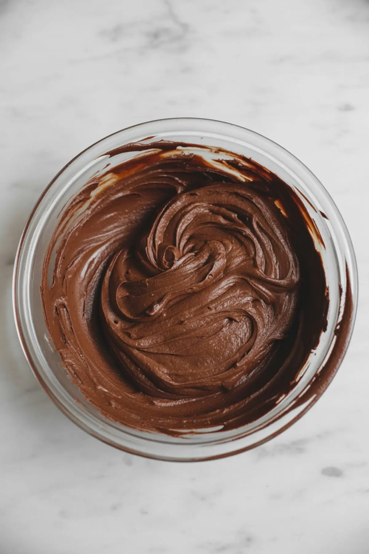 Glass bowl filled with smooth chocolate cake batter that has thick swirls and glossy texture on a marble surface.
