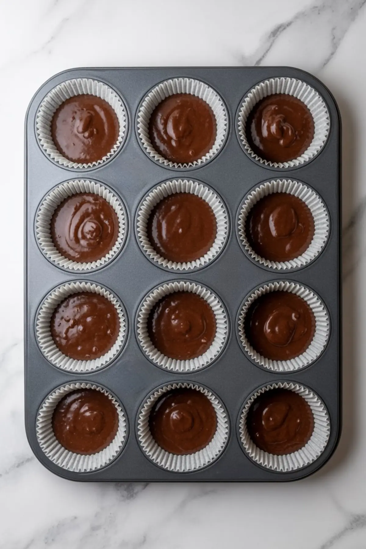 Metal muffin pan lined with paper cupcake liners filled with chocolate batter ready for baking.
