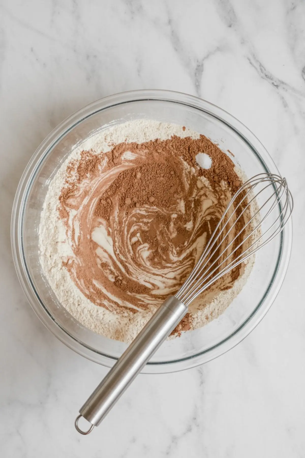 Glass bowl with partially mixed flour, cocoa powder, and dry ingredients using a metal whisk on a marble countertop.