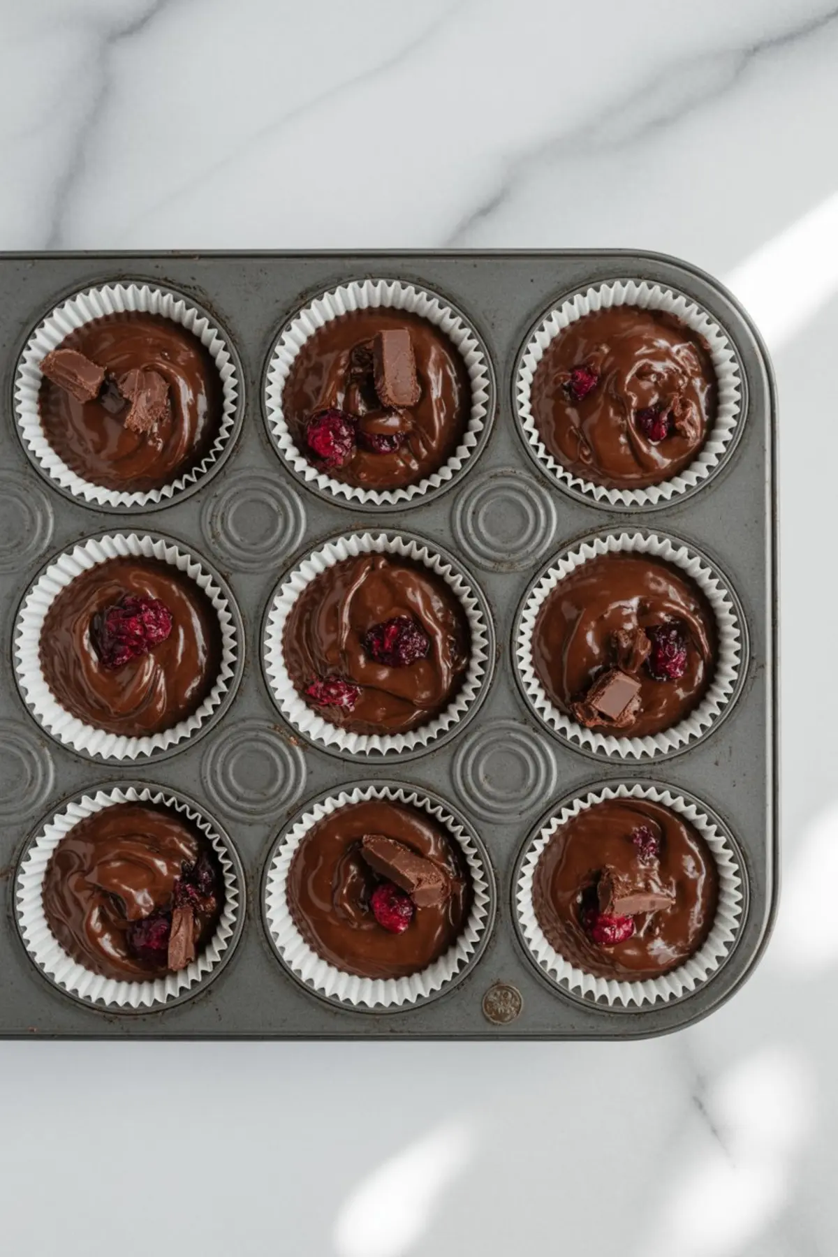 Muffin tin filled with chocolate batter in white liners, each portion topped with chunks of chocolate and cranberries, ready for baking.
