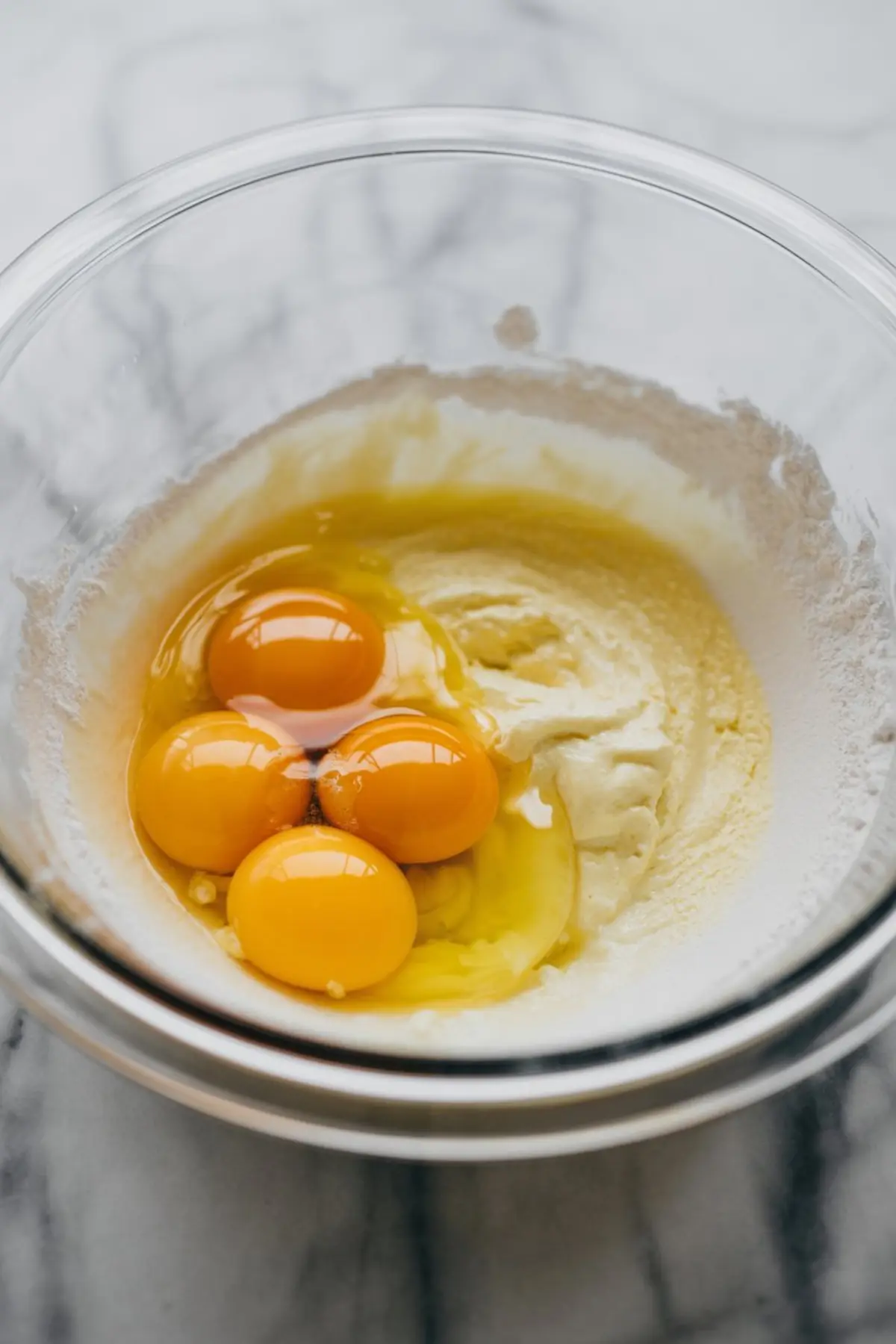 A mixing bowl containing raw egg yolks cracked into a pale batter mixture, set on a white marble surface.