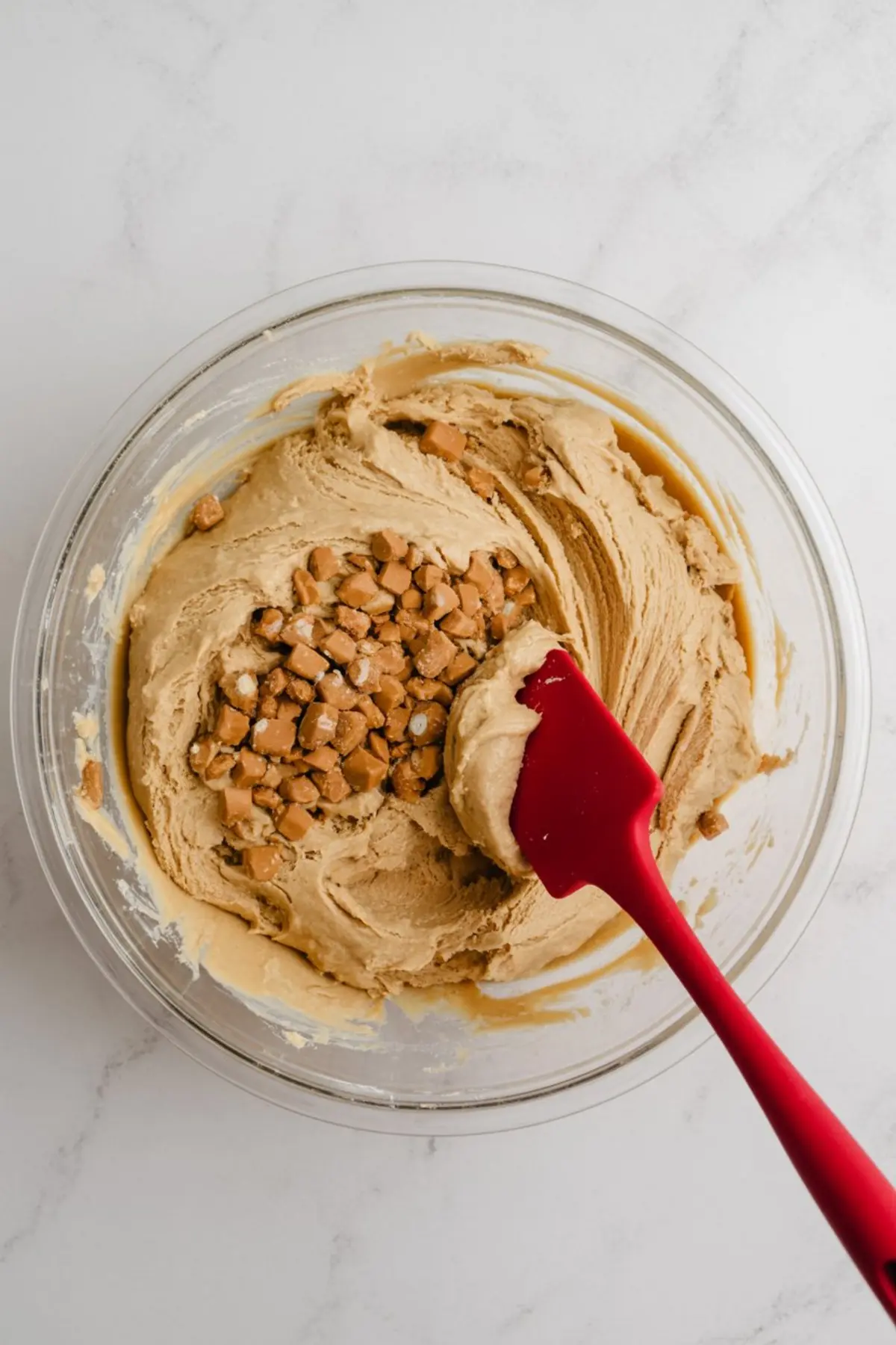 A glass bowl of creamy cookie dough with chunks of toffee pieces and a red spatula resting on the side.