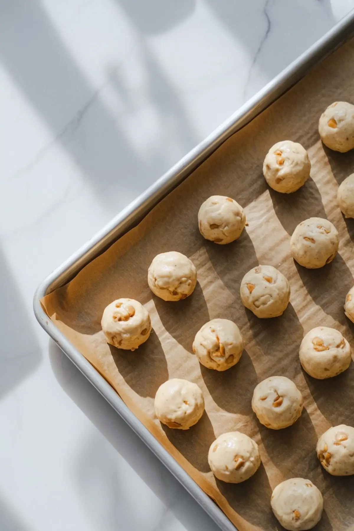 A parchment-lined baking sheet with rows of raw cookie dough balls studded with toffee bits, ready to bake under natural light on a white marble countertop.