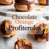 Chocolate orange profiteroles stacked on a marble cake stand, topped with glossy chocolate ganache and orange peel curls, with one cream-filled pastry cut open to show the inside.