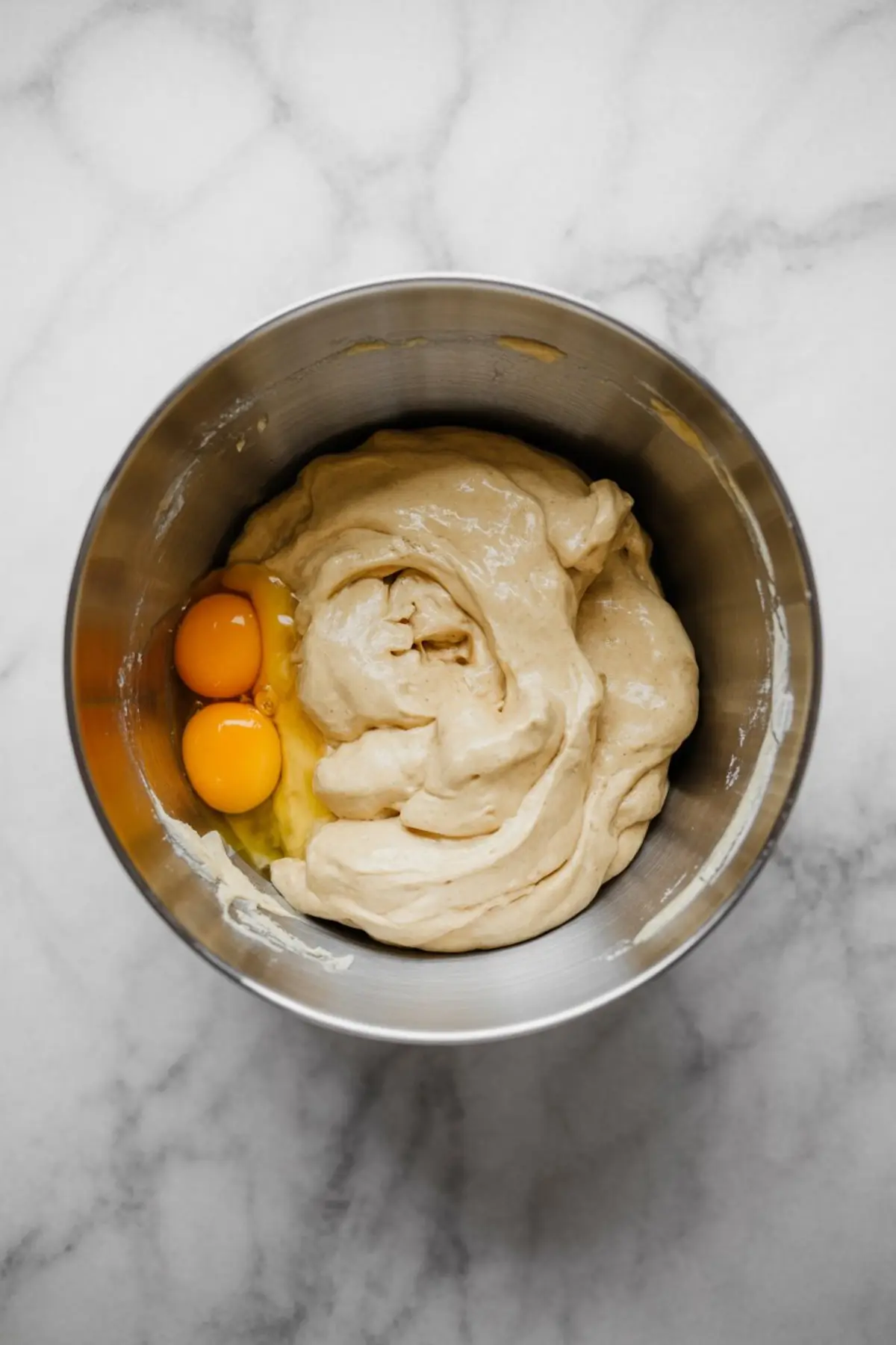 Thick dough mixture in a stainless steel mixing bowl with two cracked egg yolks resting on top, set on a marble countertop.