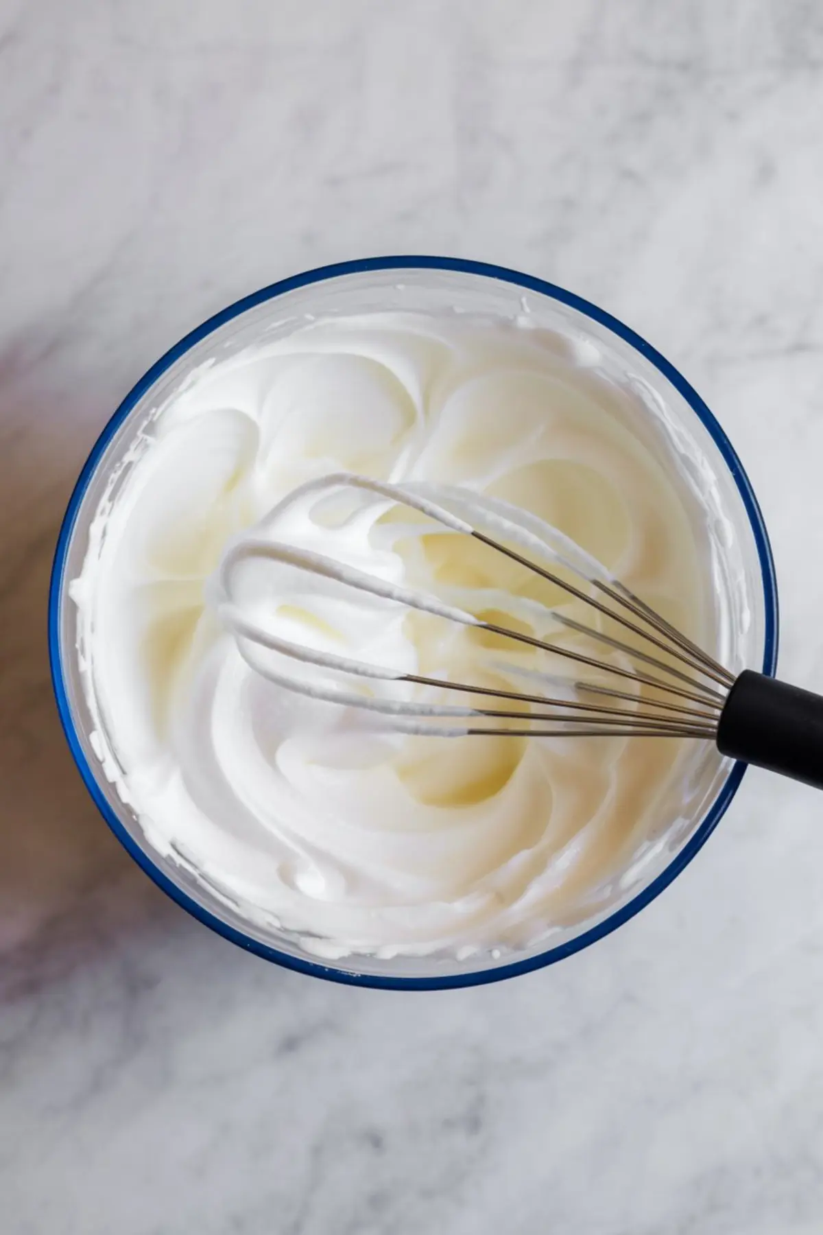 A mixing bowl filled with stiffly whipped cream, featuring smooth peaks and a metal whisk coated with fluffy white cream, set on a light marble surface.