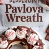 Overhead shot of a chocolate peppermint pavlova wreath decorated with piped whipped cream, peppermint candy pieces, and chocolate shards beside a text card that reads “Chocolate Peppermint Pavlova Wreath.”