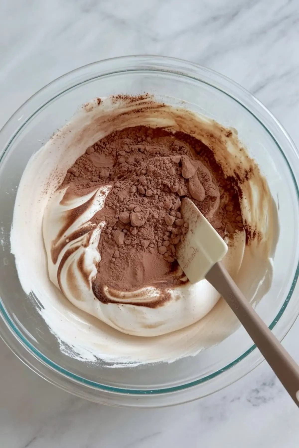 Mixing bowl filled with fluffy whipped cream and cocoa powder being folded together with a spatula to make chocolate meringue batter.