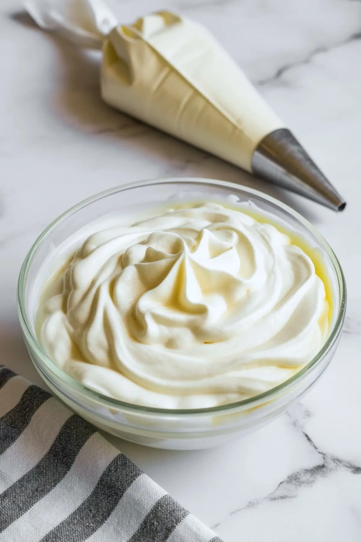 Glass bowl of smooth whipped cream with decorative swirls beside a piping bag on a marble countertop, ready for dessert decorating.