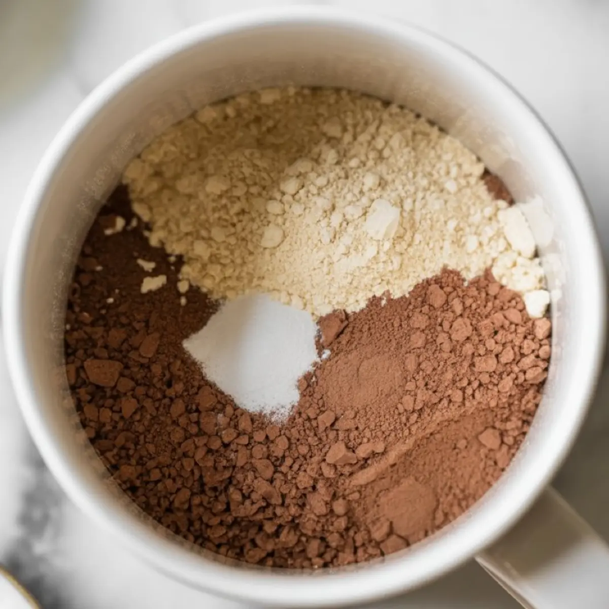 Overhead view of a white mug filled with dry ingredients for a chocolate protein mug cake, including cocoa powder, protein powder, and baking soda, arranged in neat sections before mixing.
