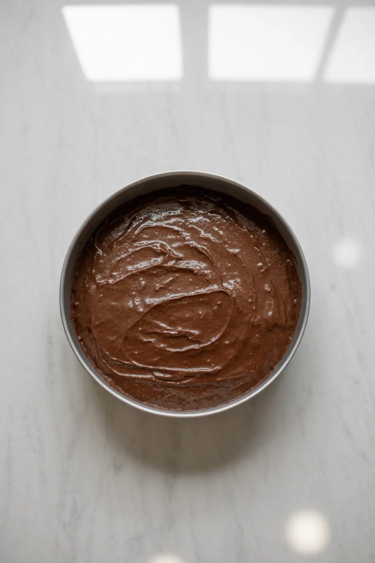 Smooth, dark chocolate cake batter swirled inside a round cake pan, ready for baking, sitting on a white marble surface with overhead light reflections.